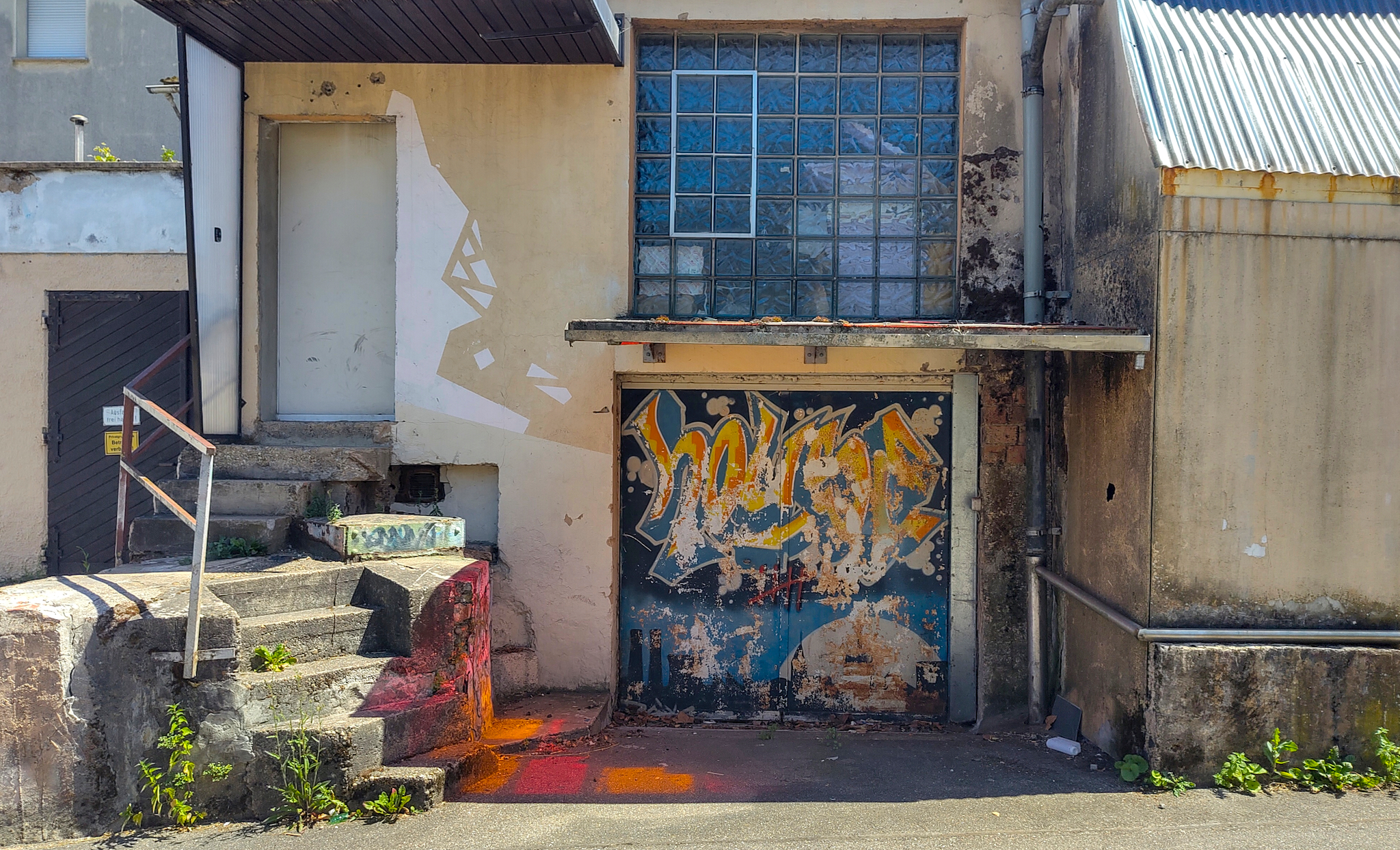 Dieses Bild zeigt einen verschachtelten Gebäudekomplex, der Graffiti auf Wänden und Garagentor aufweist. Das Gebäude ist durch verblasste Farbe, Rost und Risse in der Fassade gekennzeichnet. Das Sonnenlicht scheint durch ein mit farbigen Glasscheiben gestaltetes Garagenvordach.